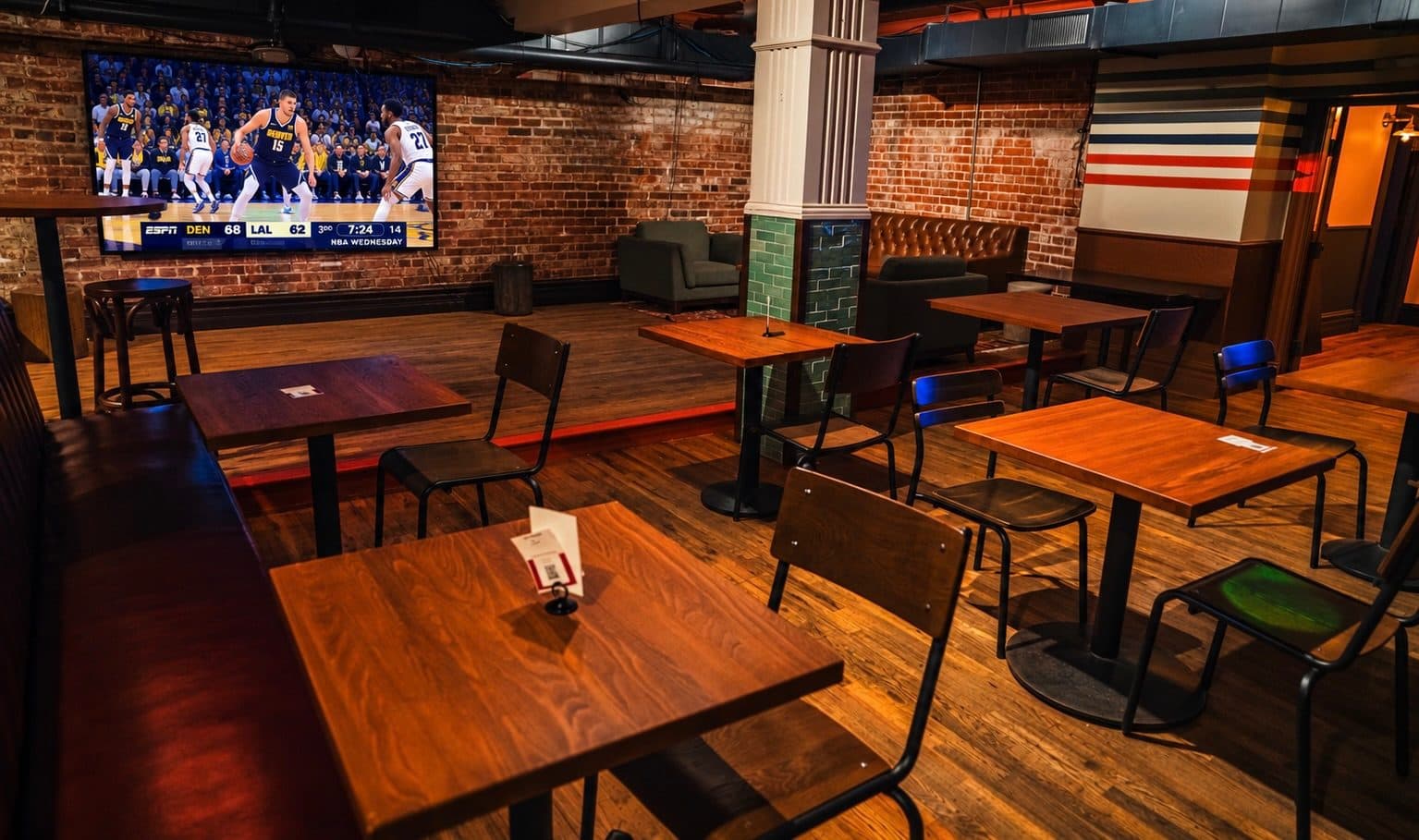 Tables and lounge chairs under exposed brick, a basketball game on the big screen, at BARna